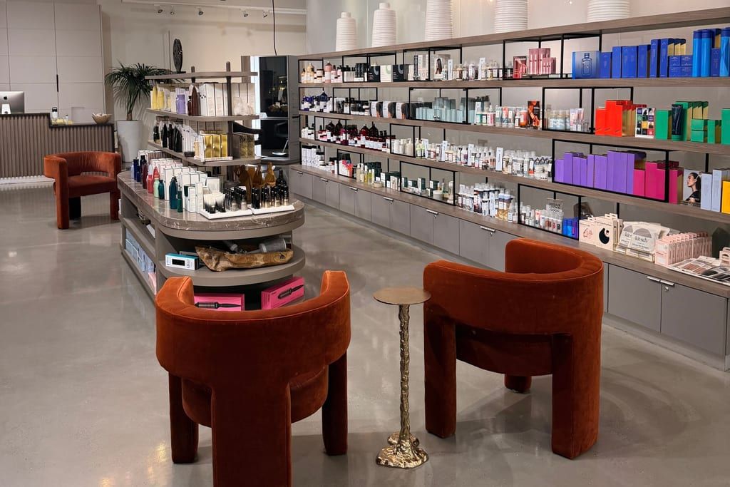 Square Salon's retail floor: a wall of open shelving stocked with hair and skin care, a marble product island, two rust velvet armchairs around a brass side table, and polished concrete underfoot.
